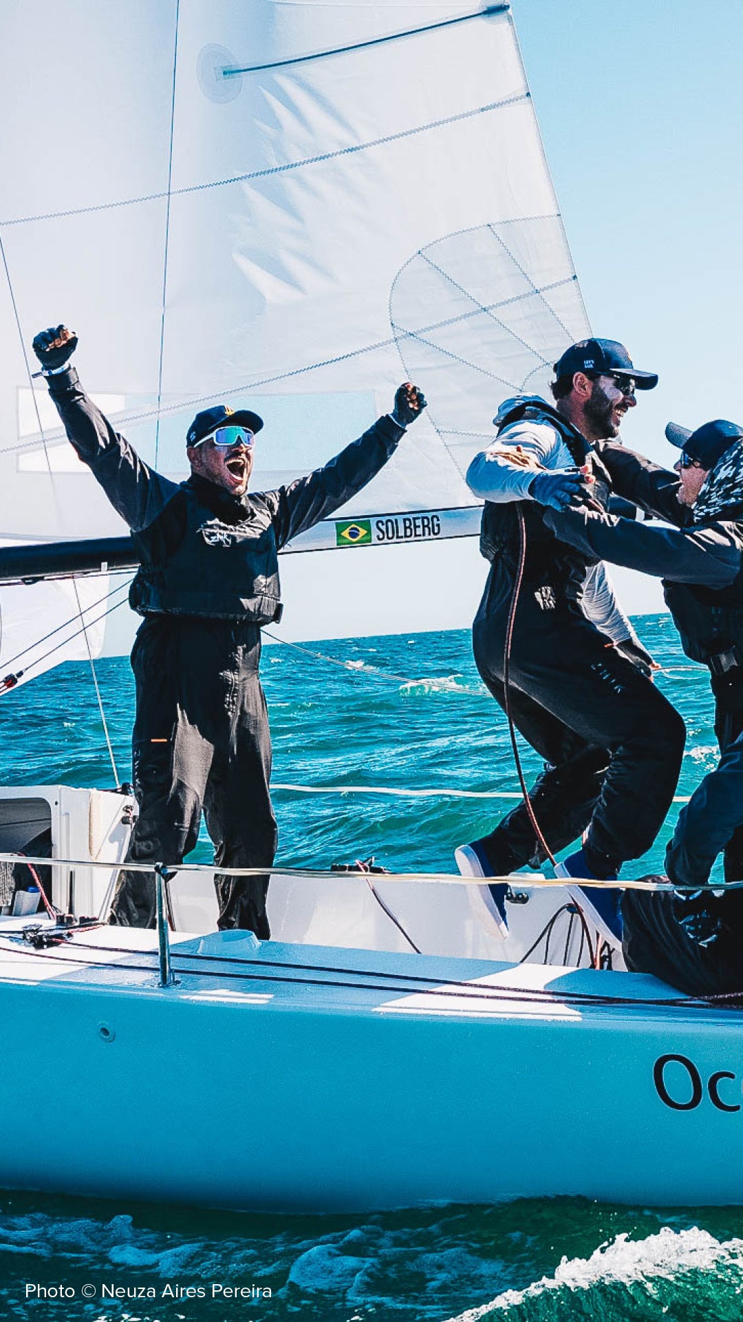 A white J/70 sailboat racing on blue ocean waters under a clear sky, with four people aboard cheering and balancing on the deck.